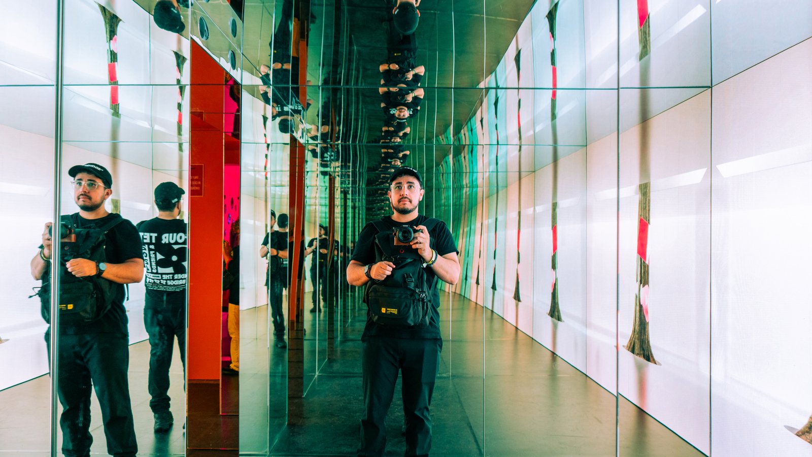 Photograph of Alex taken by himself, holding his camera just below his face, standing in a room full of mirrors so that you can see many reflections of him from the front and behind on all sides on the room.